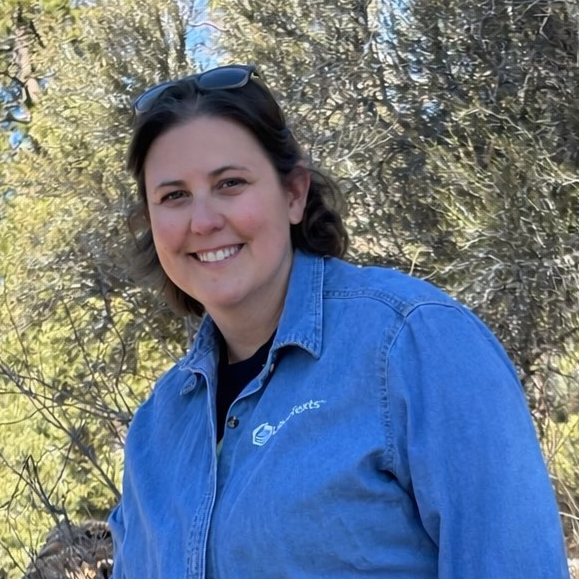 Woman in a blue shirt taking a selfie on a beach with the forest in the background  